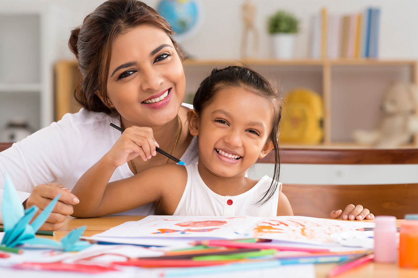 Picture of a Hispanic teacher and kindergarten-aged girl smiling; the girl is holding a paintbrush and is painting