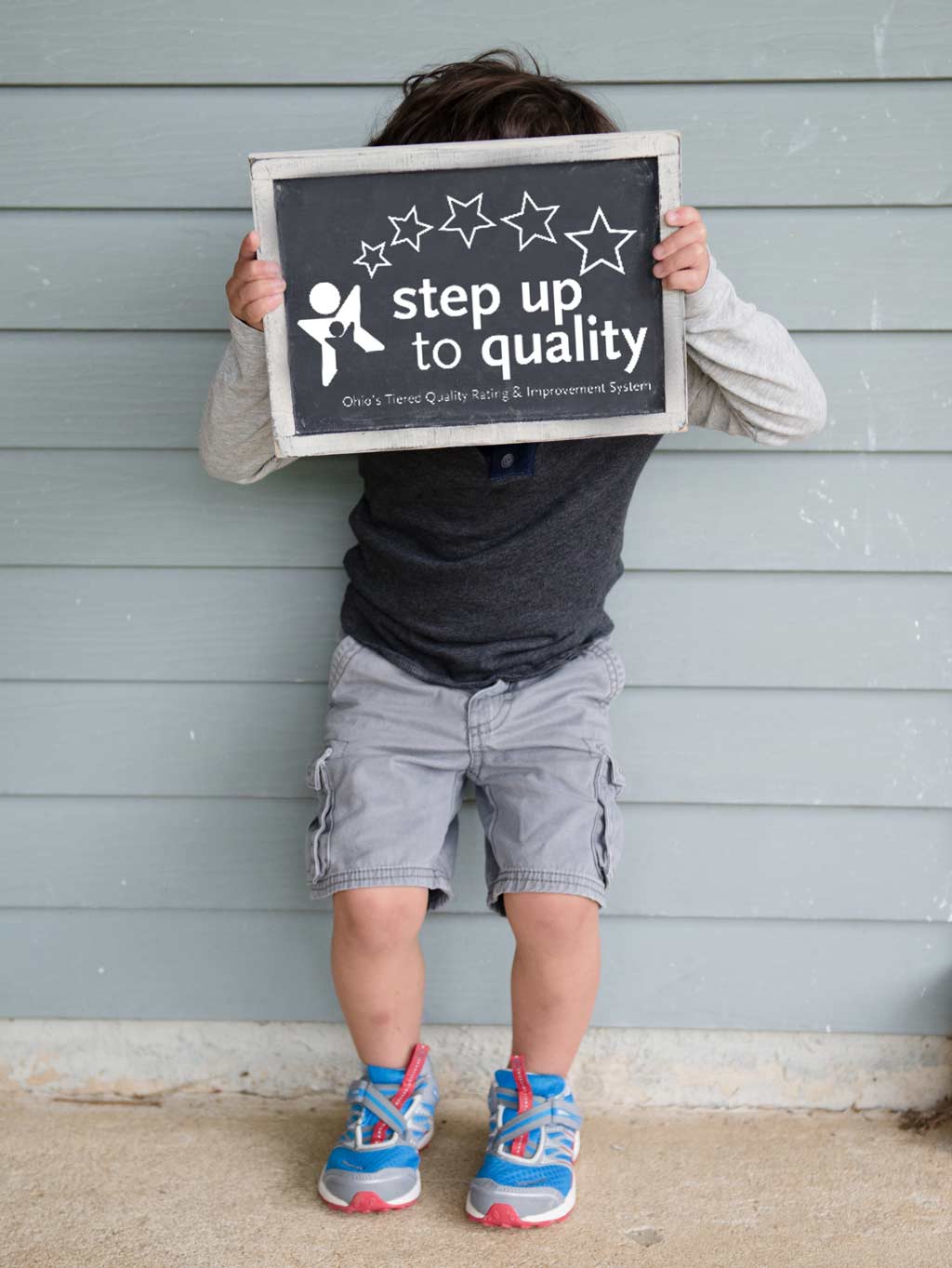 Picture of a dark-haired kindergarten-aged boy holding a small chalkboard in front of his face; the chalkboard has a graphic on it that says: 'Step up to quality'
