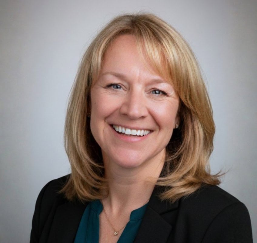 A woman with shoulder-length blonde hair smiles, wearing a black blazer over a teal top, posed against a plain light gray background.