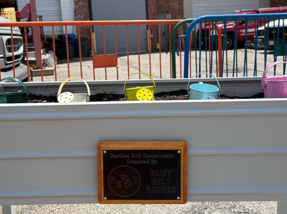 A metal planter box with small colorful watering cans sits outdoors. A plaque reads “Garden Bed Generously Donated By Rust Belt Riders.”.