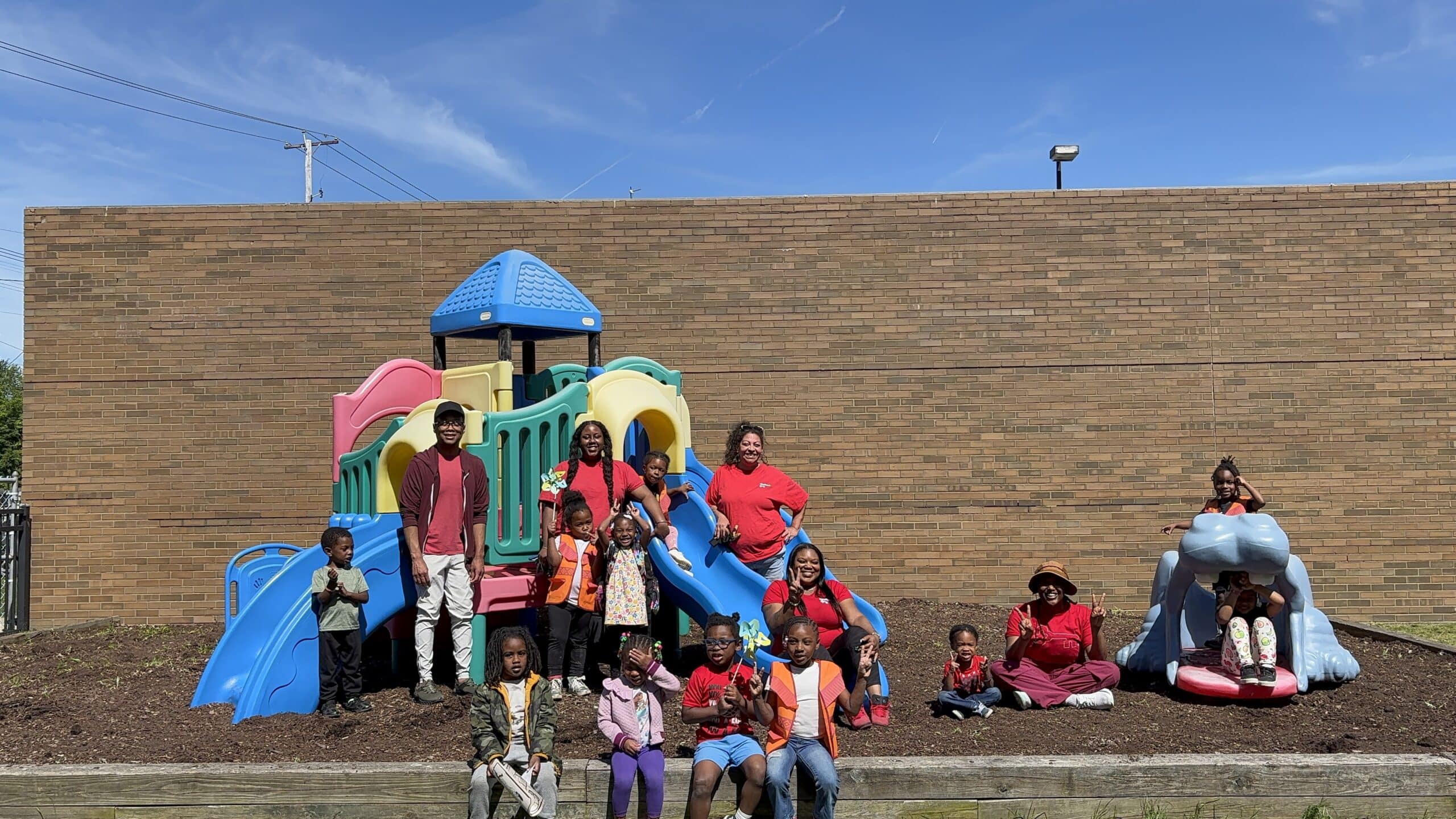 A group of children and adults, some wearing red shirts, pose in front of a colorful playground slide structure with a brick wall in the background on a sunny day.