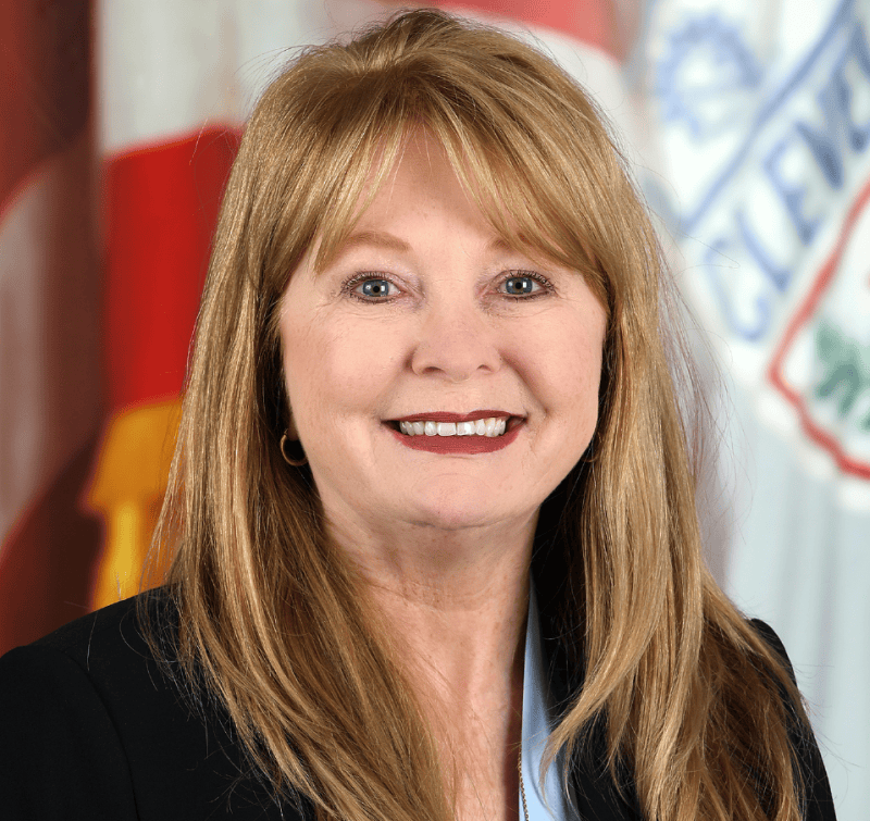 A woman with long blonde hair smiles at the camera, wearing a black blazer over a light blue top. An American flag and another flag are visible in the background.
