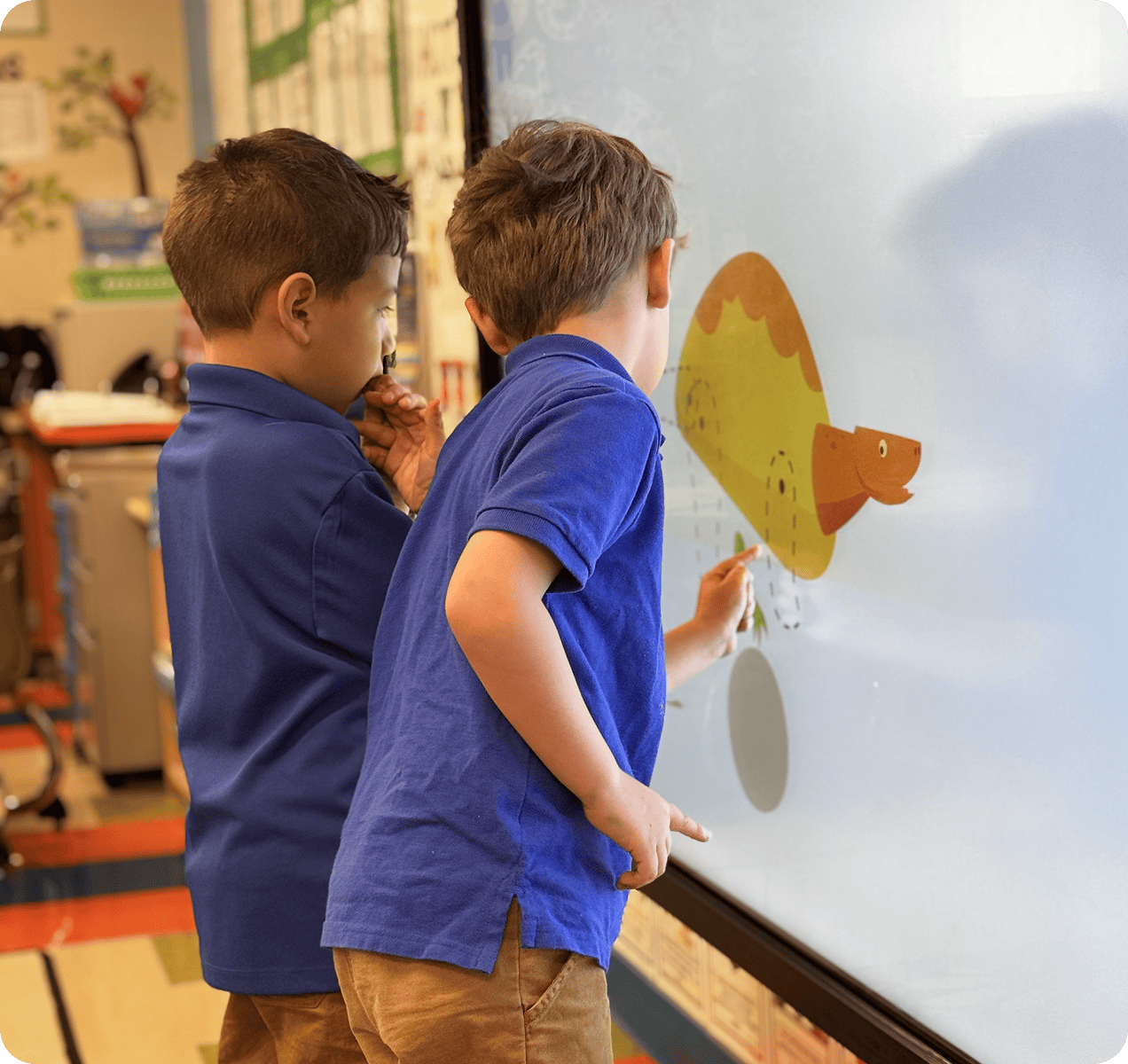 about-1 Two young boys in blue shirts use a digital whiteboard to interact with an illustrated dinosaur in a classroom setting.