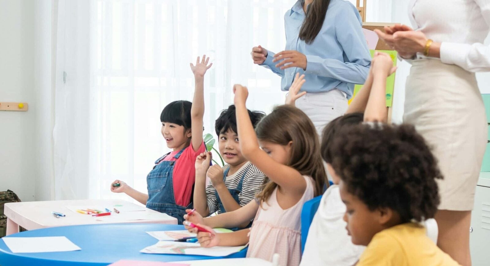 A group of young children sit at tables in a classroom, some raising their hands, while two adults stand nearby. Art supplies and papers are on the tables.