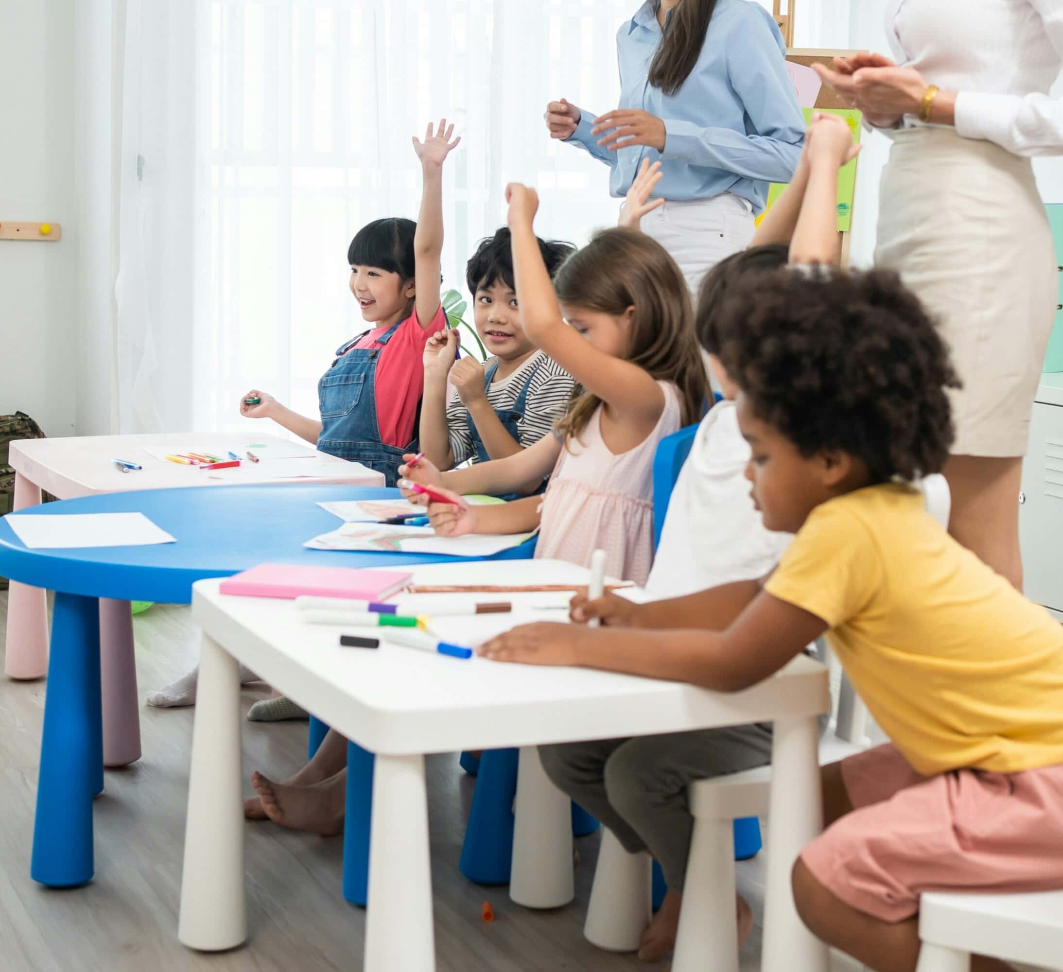 A group of young children sit at tables in a classroom, some raising their hands, while two adults stand nearby. Art supplies and papers are on the tables.