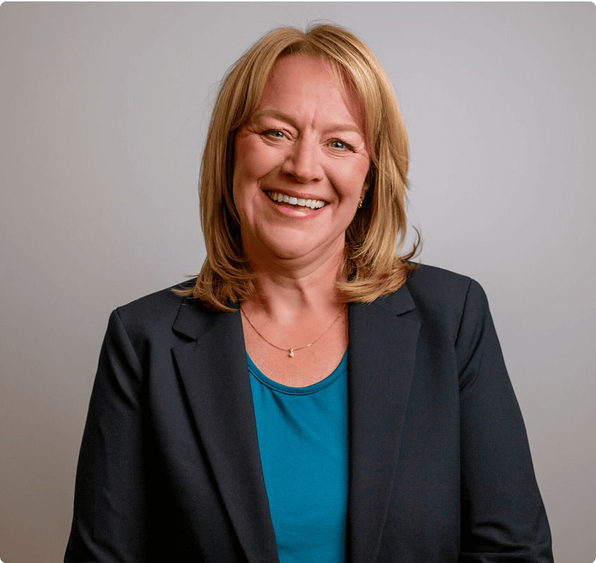 A woman with blonde hair wearing a black blazer and blue top smiles in front of a plain light gray background.