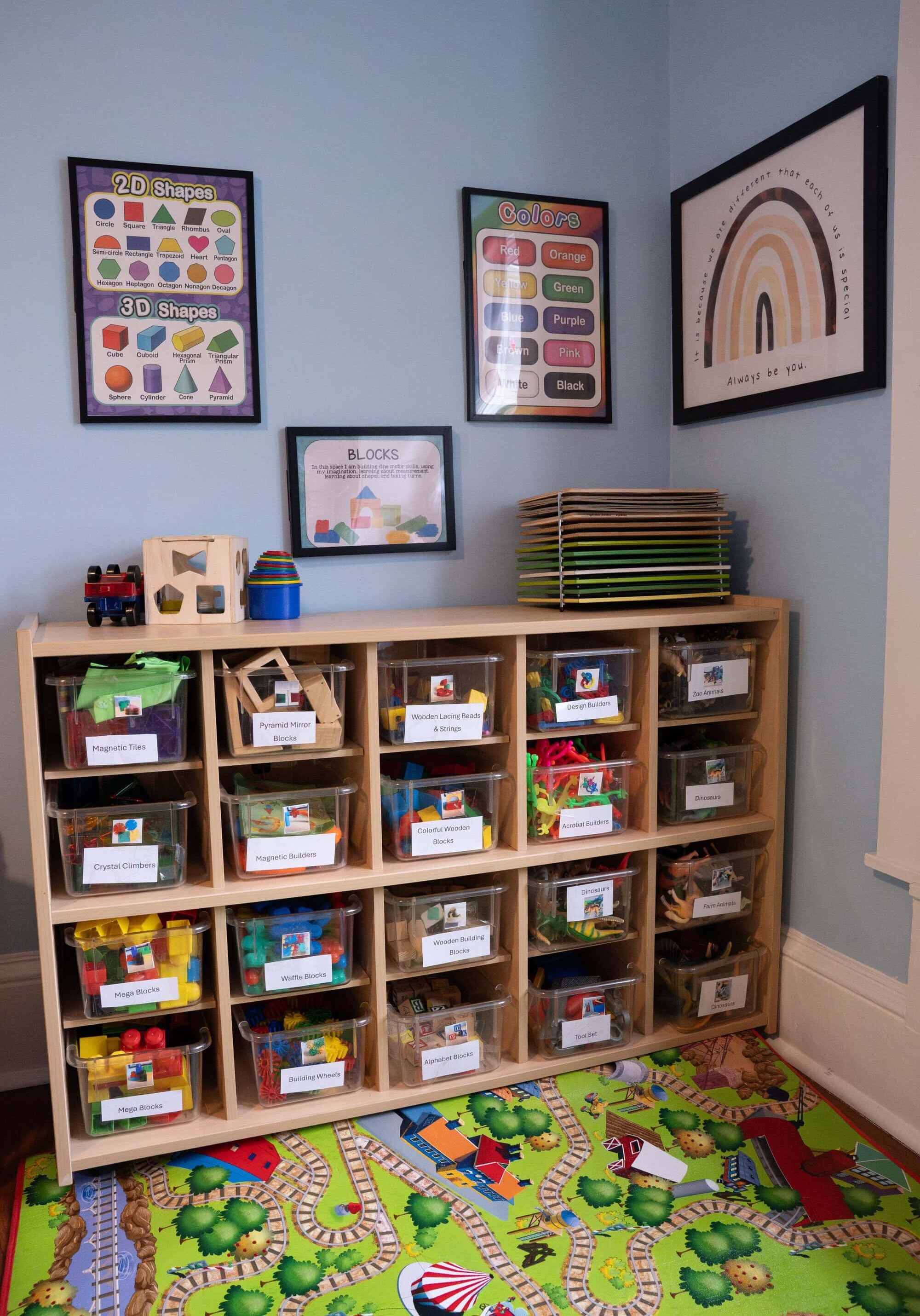 A wooden shelf holds labeled bins of toys and blocks in a playroom with educational posters on blue walls and a colorful play mat on the floor.