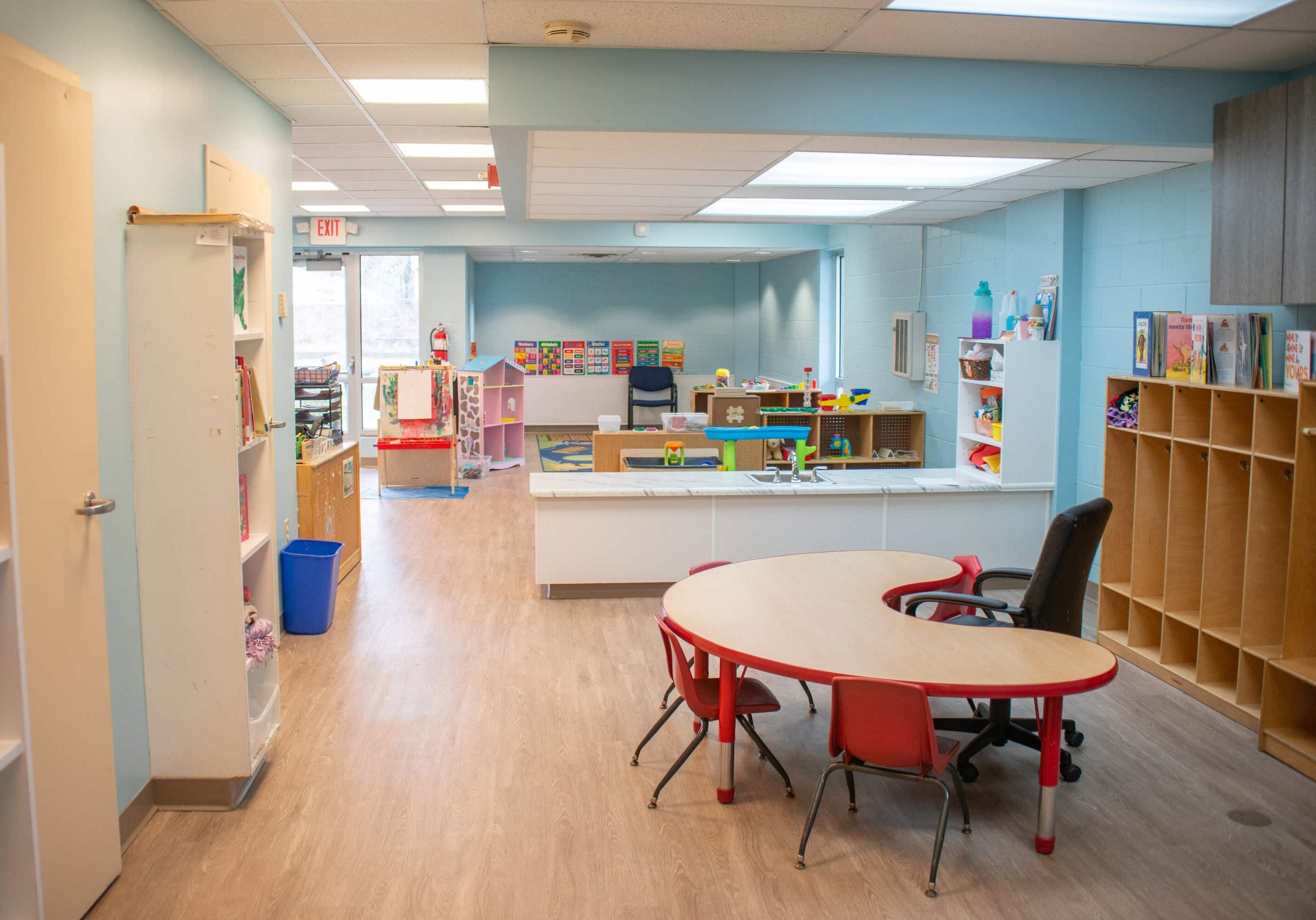 A brightly lit preschool classroom with shelves, toys, art supplies, and small tables with red chairs on a wood floor.