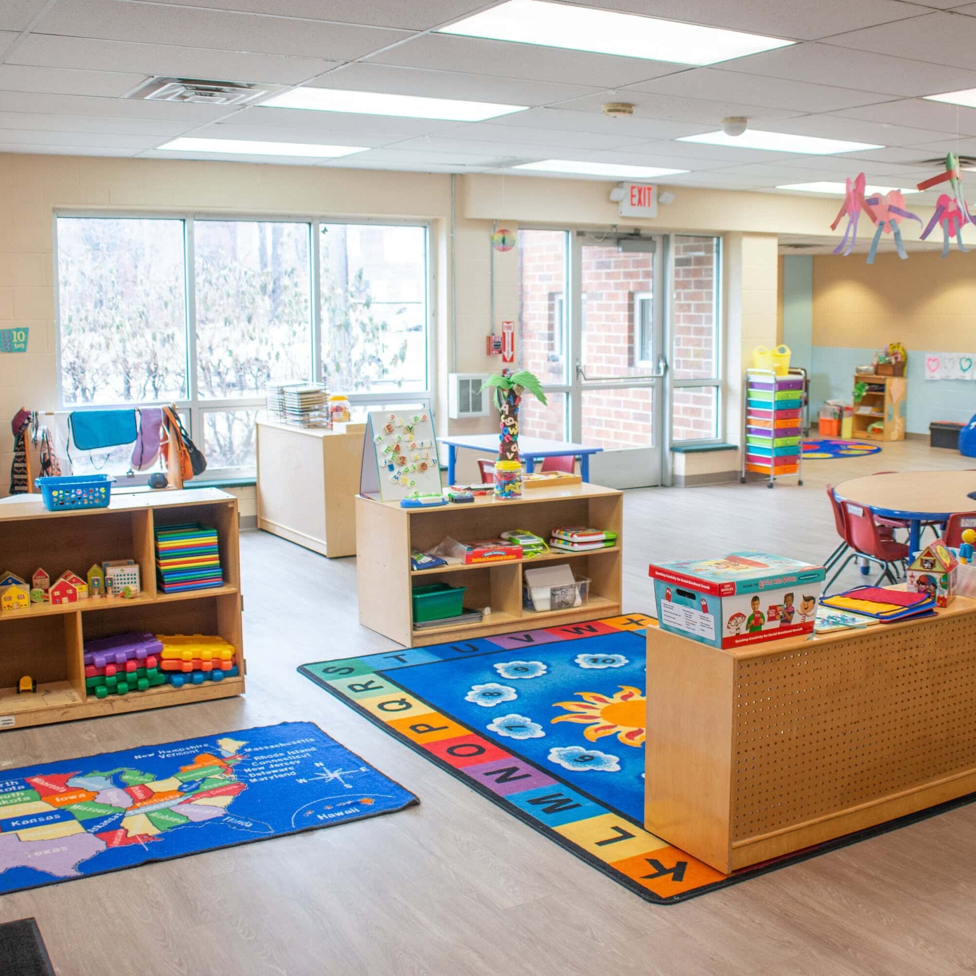 A bright daycare classroom with organized shelves of toys, colorful rugs, child-sized furniture, large windows, and educational decorations.