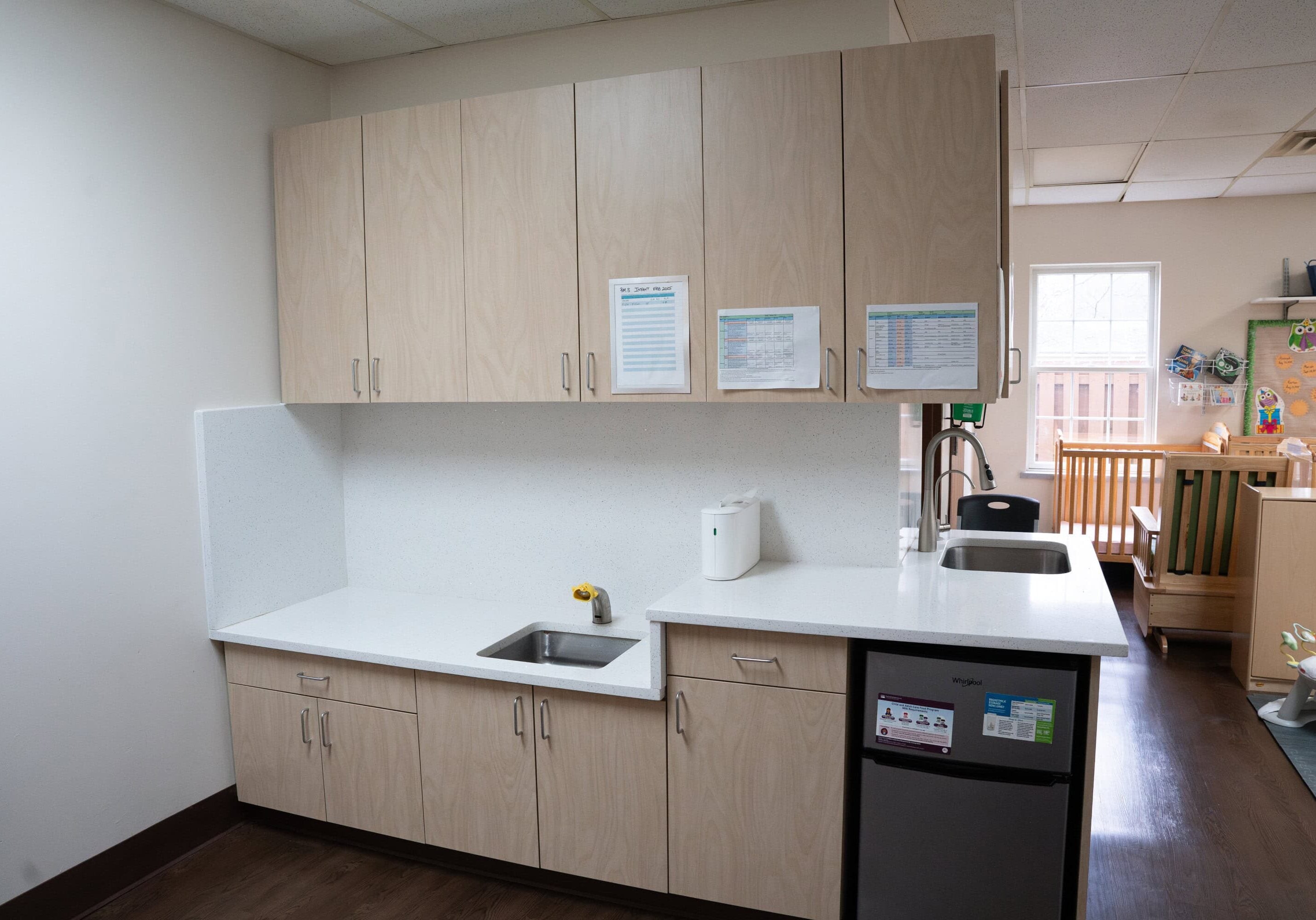 A small kitchenette with light wood cabinets, a sink, a mini fridge, paper towels, and notices on the wall, next to a play area with a crib.
