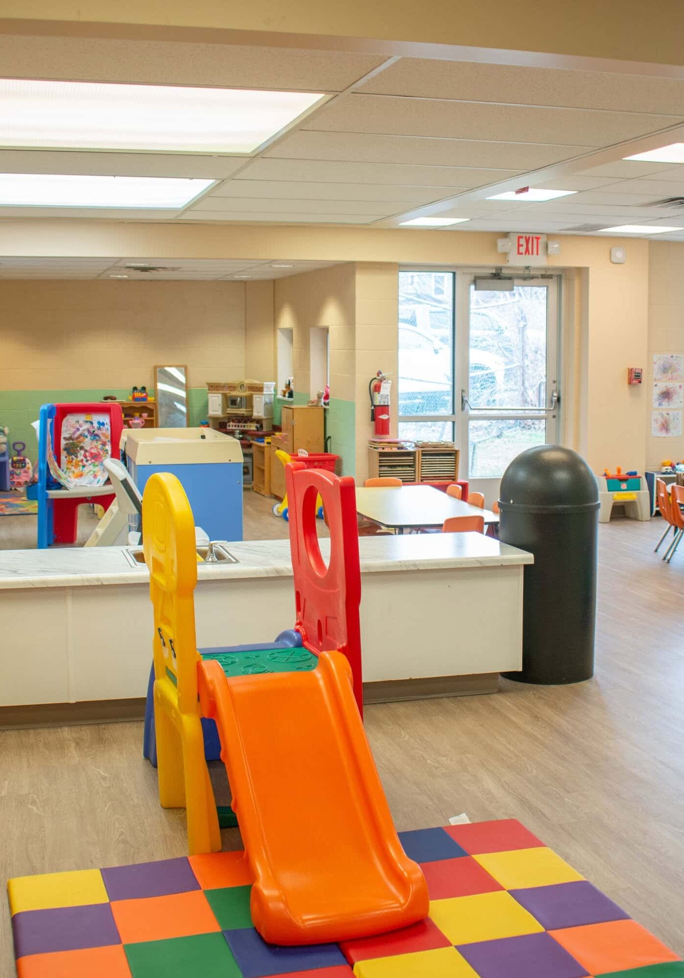 A brightly lit daycare classroom with colorful play equipment, a slide, toys, tables, and chairs on a wood floor, with large windows in the background.