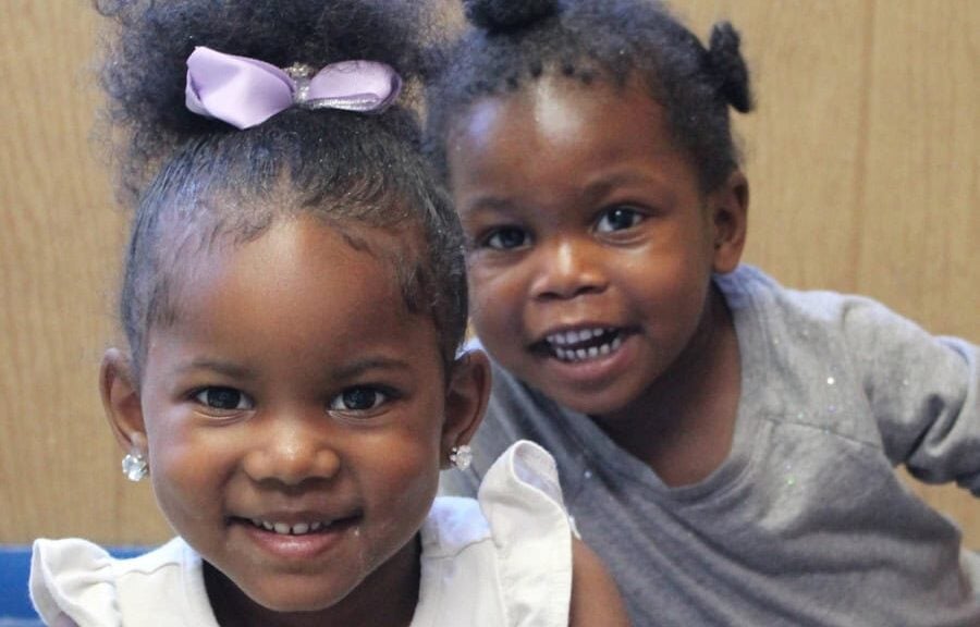 Two young girls smile at the camera; one wears a white top and has a purple bow in her hair, while the other wears a gray shirt and stands slightly behind her.