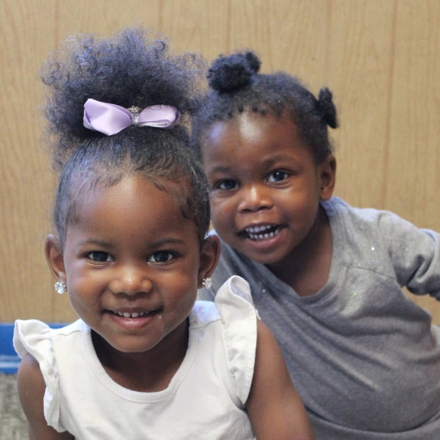 Two young girls smile at the camera; one wears a white top and has a purple bow in her hair, while the other wears a gray shirt and stands slightly behind her.