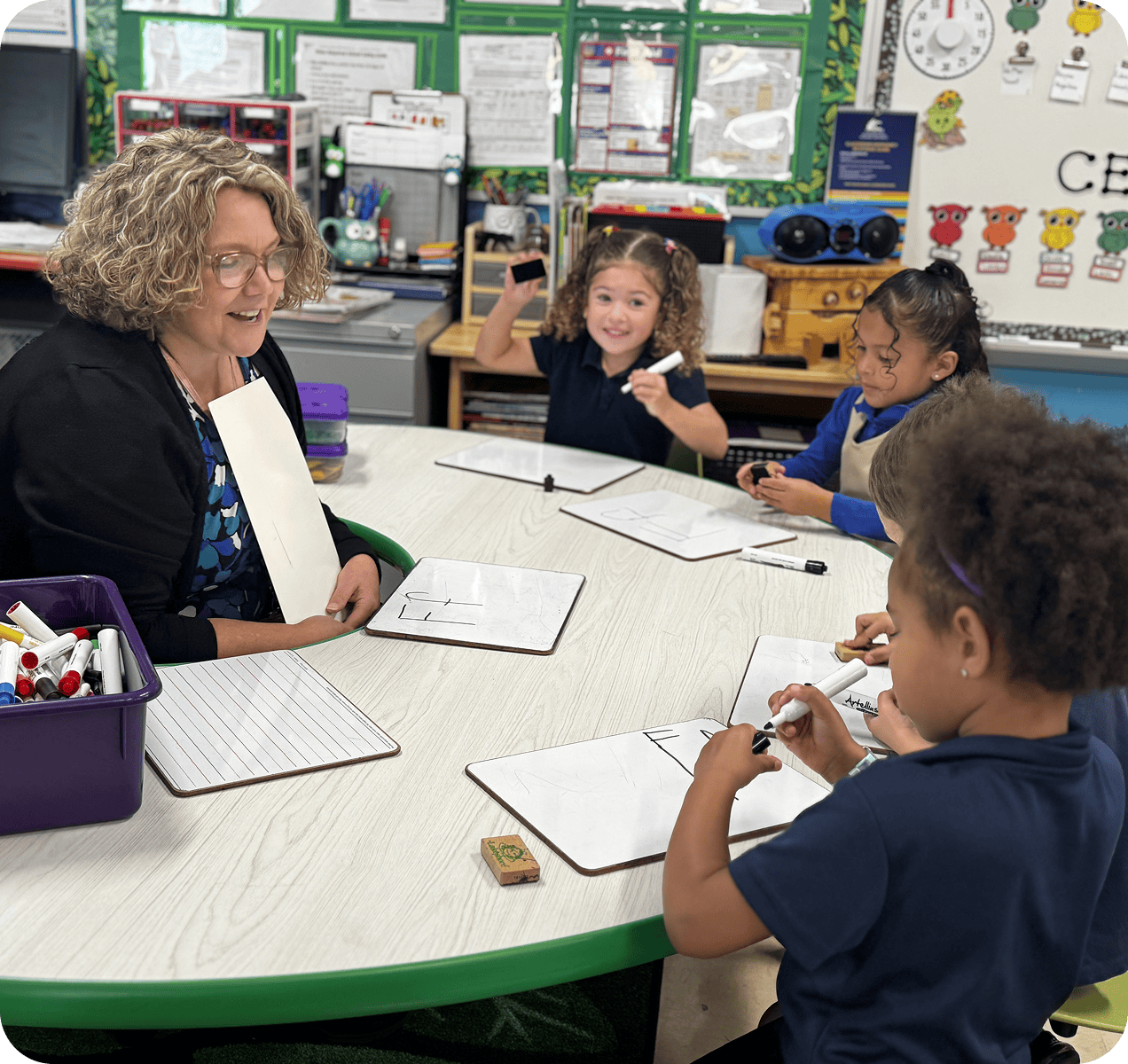 A teacher sits at a table with four young students holding whiteboards and markers, engaging in a classroom activity.