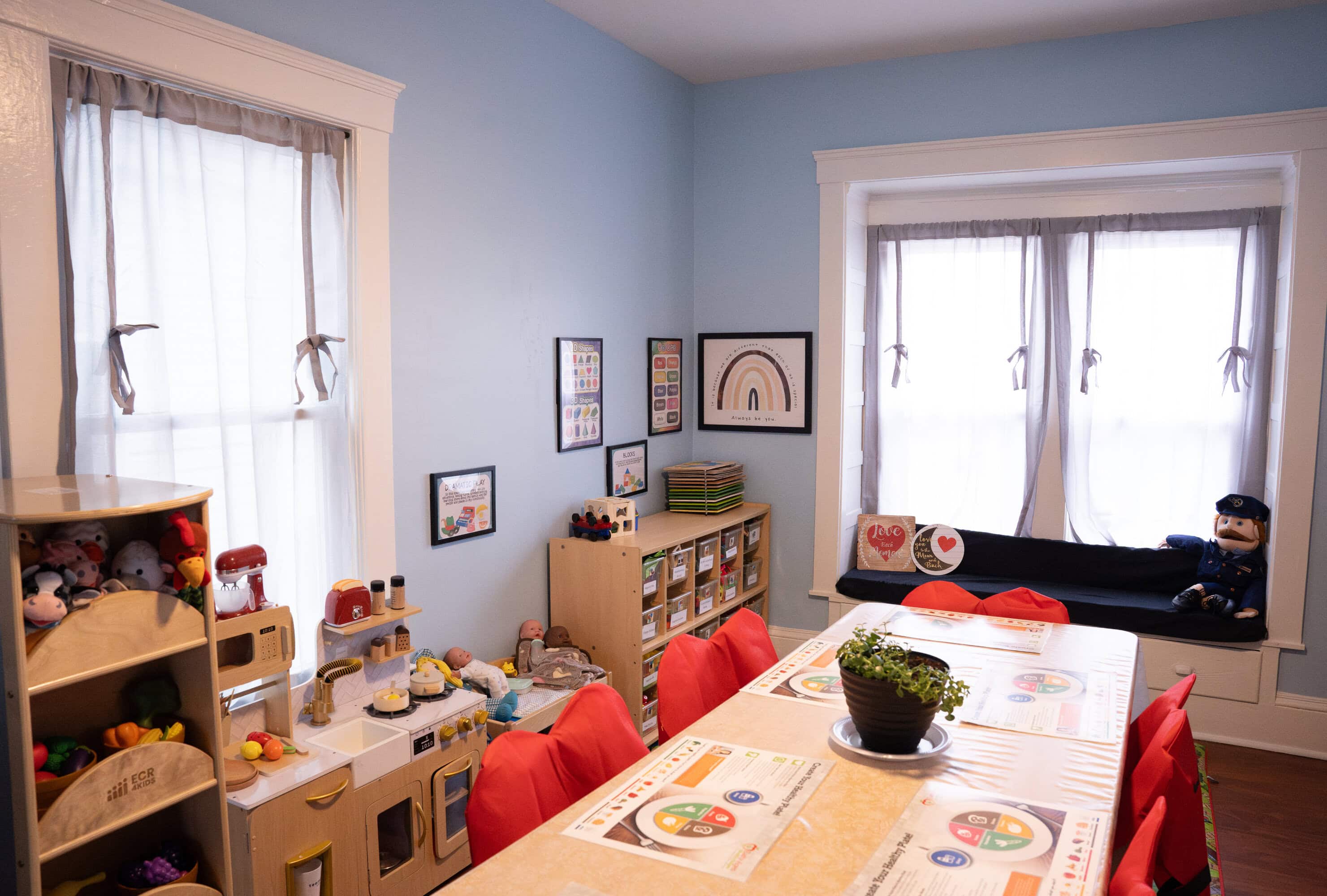 A classroom with tables and chairs, educational posters, shelves with toys and books, a play kitchen, and a window seat with a stuffed doll.