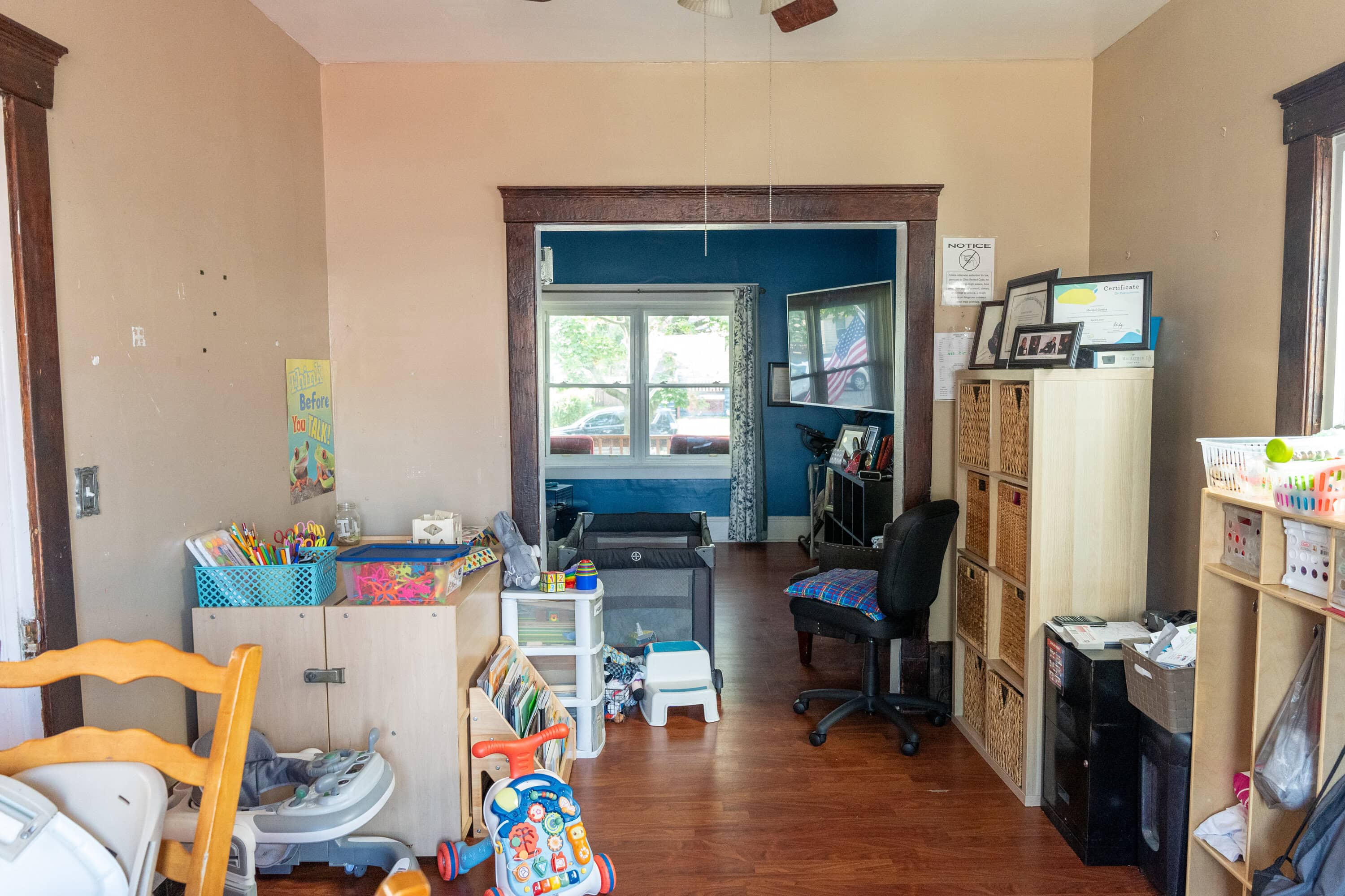 A cluttered home office and playroom with shelves, toys, a desk with a computer, and various supplies, viewed from an adjacent room with a wooden floor.