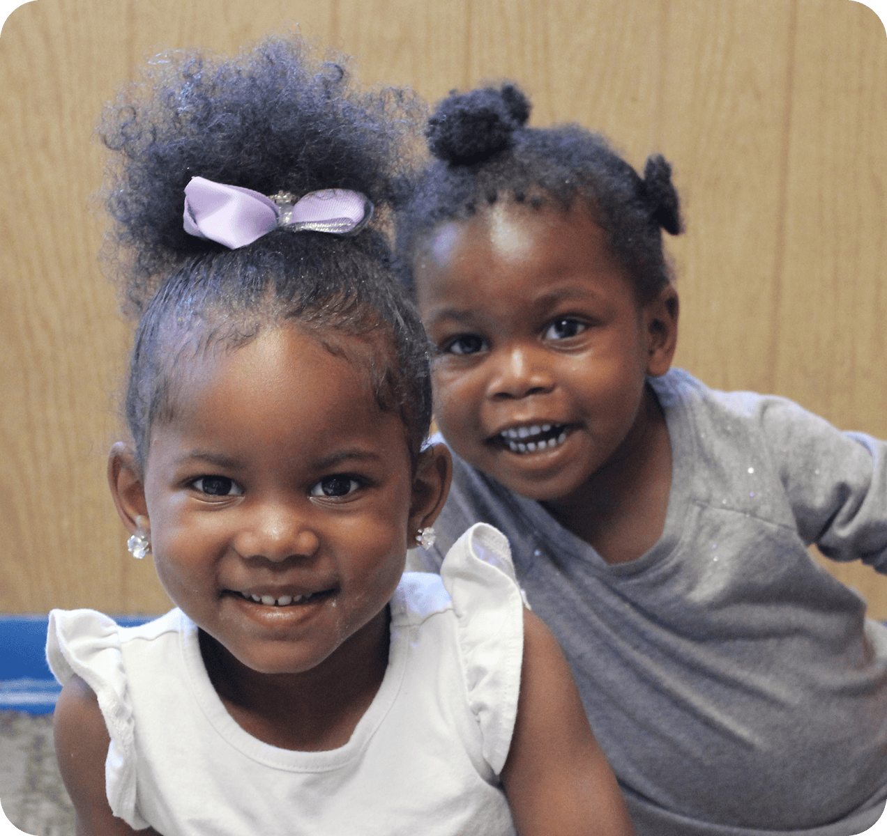 donate Two young children smiling; one wears a white top and a purple bow, while the other wears a gray shirt. They are indoors with a wooden wall background.