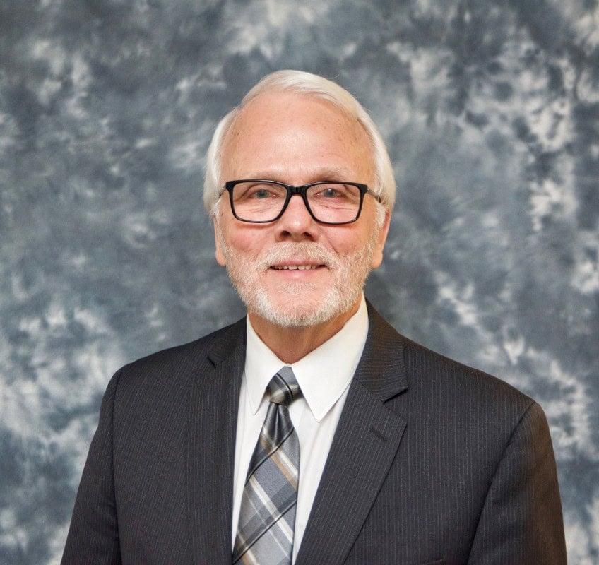 An older man with white hair and a beard, wearing glasses, a dark suit, white shirt, and plaid tie, poses in front of a mottled gray backdrop.