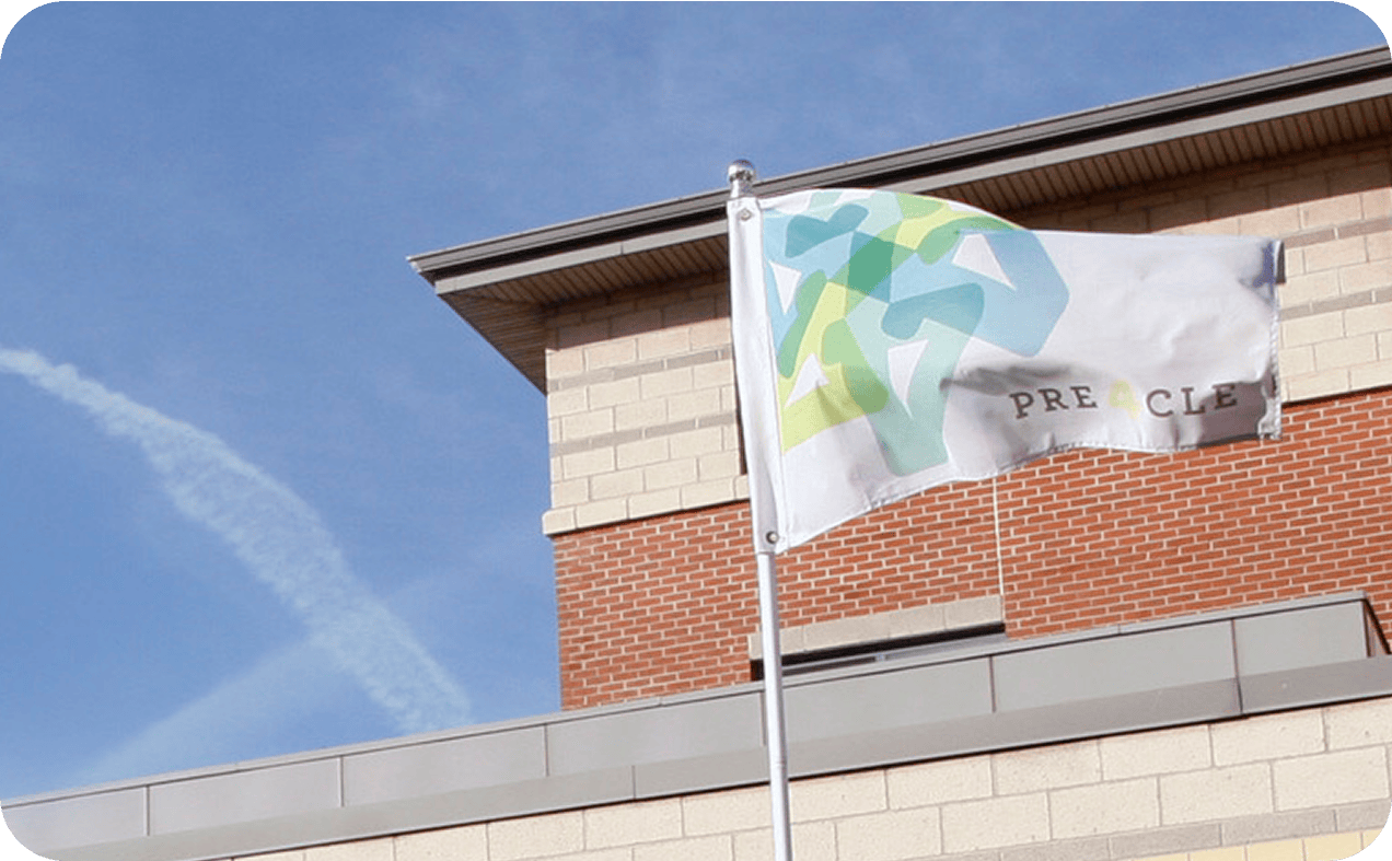 flag A white flag with green and blue abstract shapes and partial text "PRE CLE" flies in front of a brick and beige building under a blue sky.