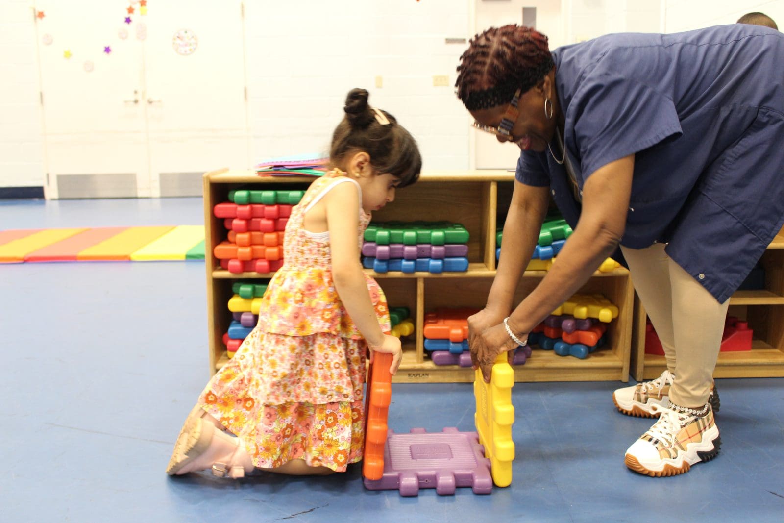 A young girl and an adult assemble colorful plastic blocks on the floor in a classroom setting.