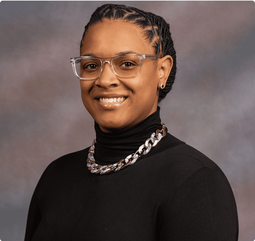 A person wearing clear glasses, a black turtleneck, and a chunky chain necklace smiles at the camera against a neutral, blurred background.