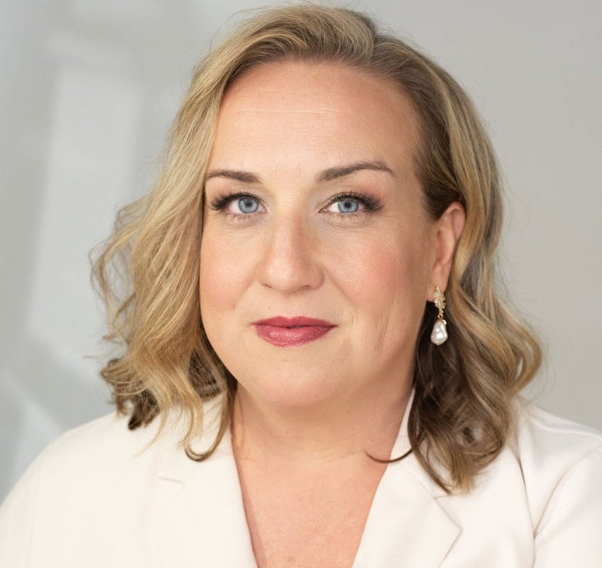 A woman with shoulder-length blonde hair, blue eyes, and light makeup, wearing a cream blazer and a dangling pearl earring, looks at the camera against a plain background.