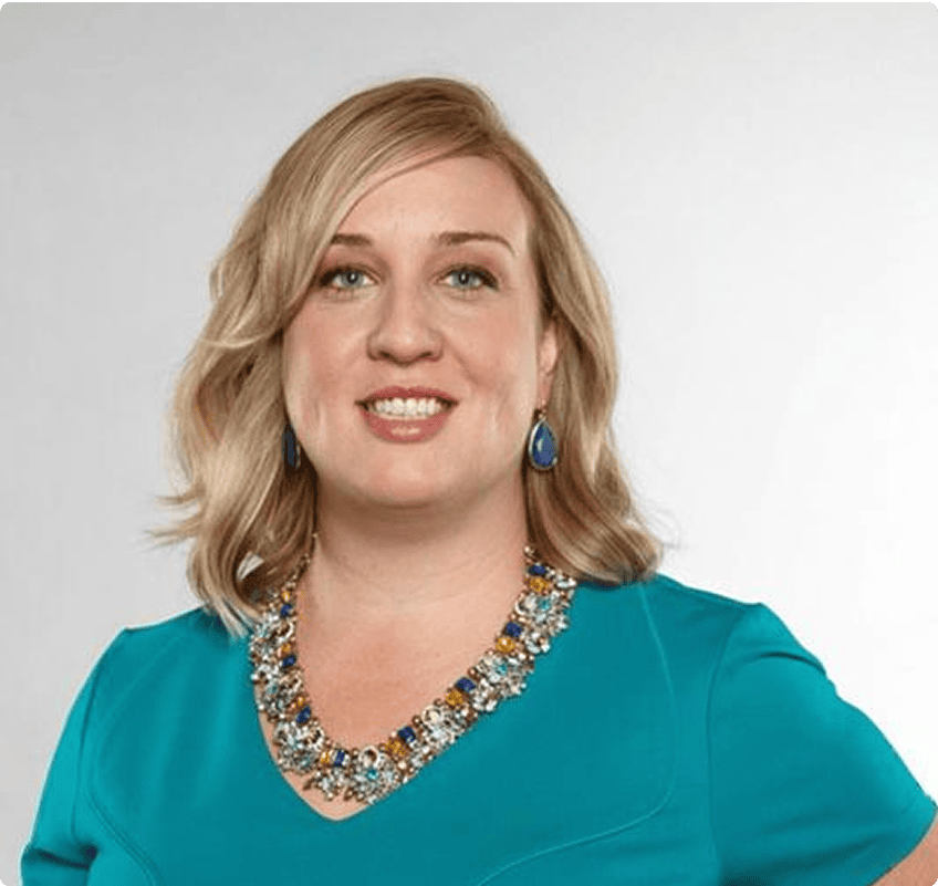A woman with blonde hair wearing a teal dress, a statement necklace, and blue earrings poses against a plain light background.