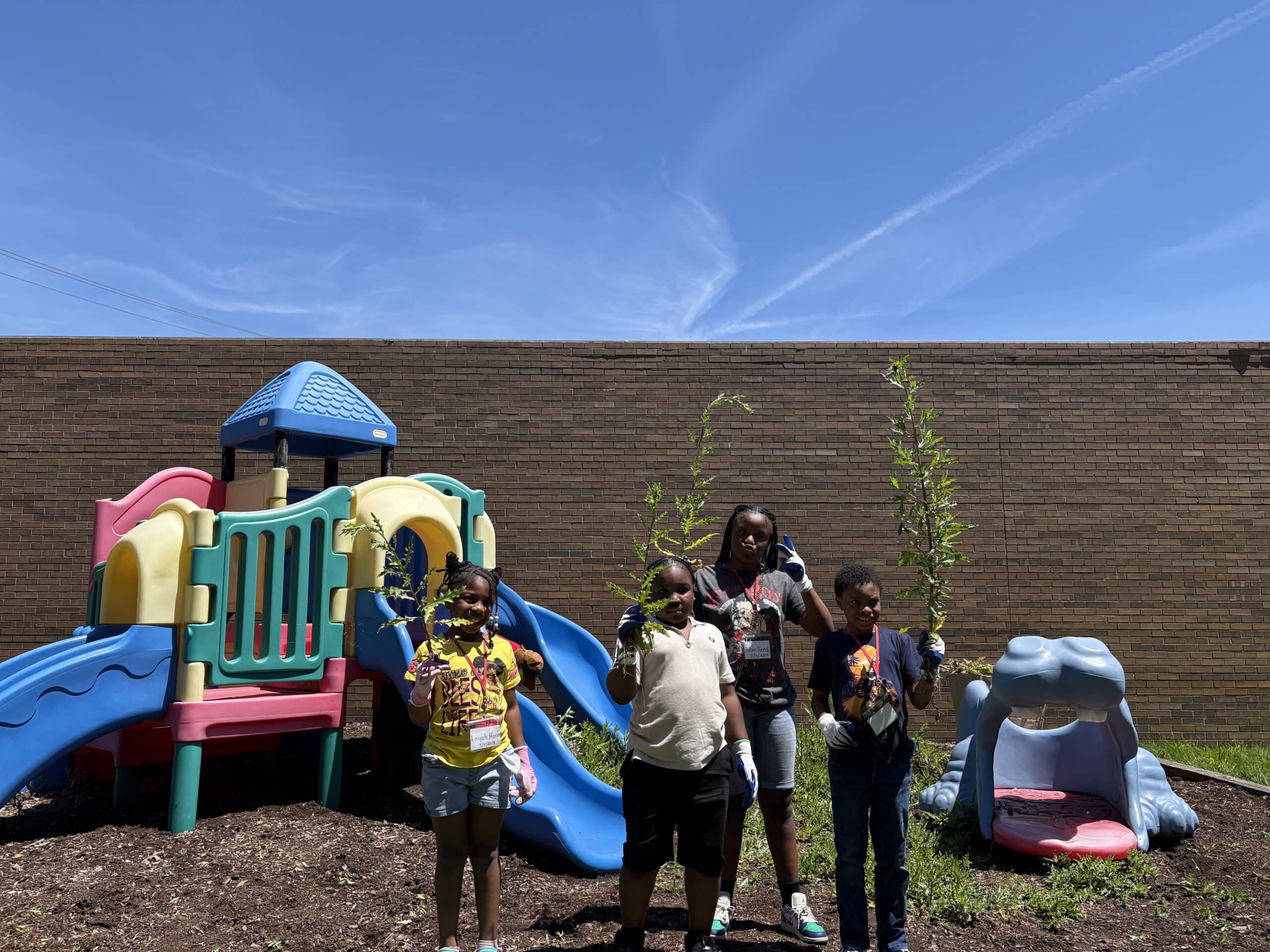 Four children stand in front of a playground slide and blue hippo structure, holding plants, with a brick wall and clear sky in the background.