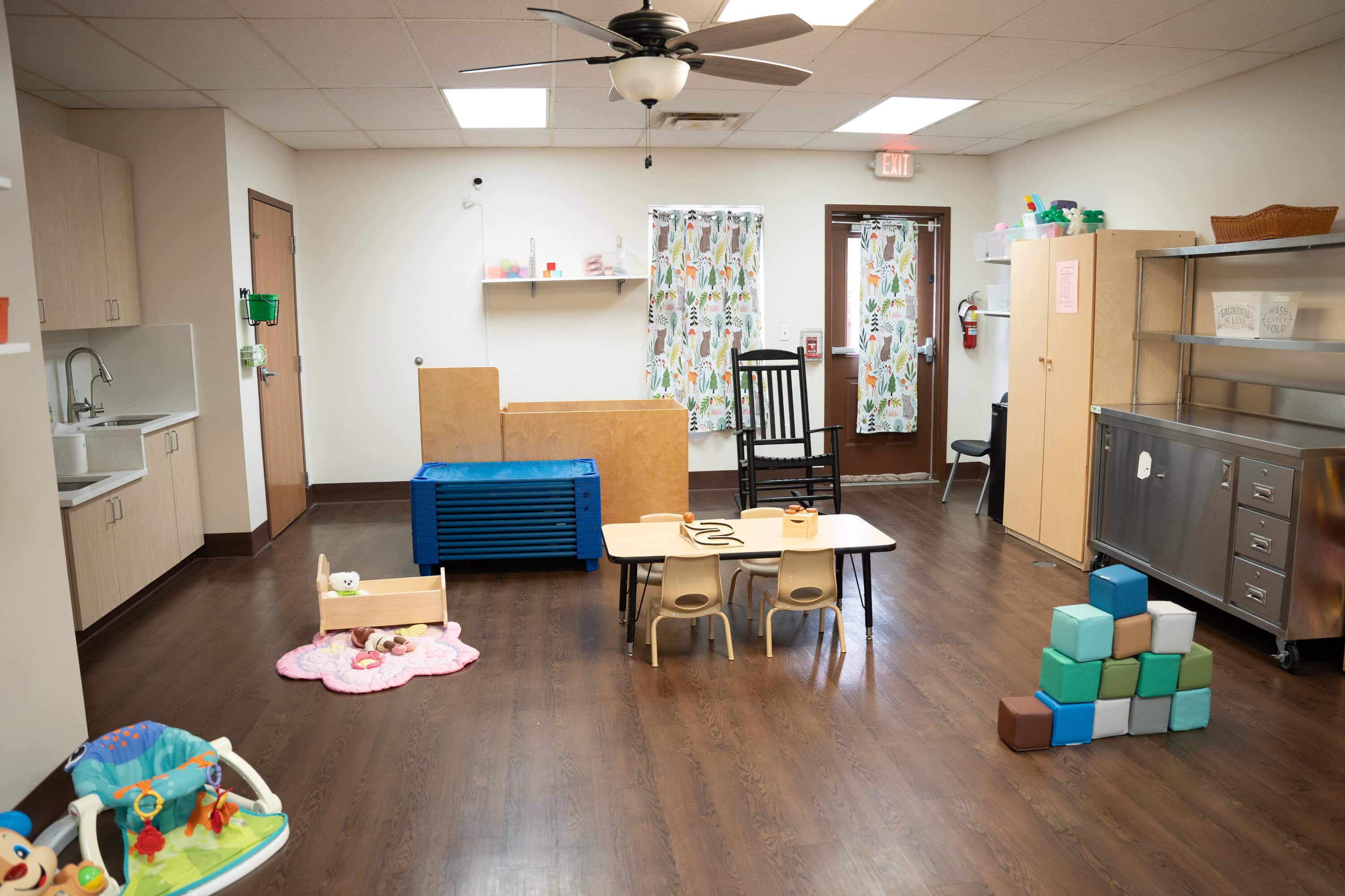 A daycare classroom with wooden floors, small tables and chairs, toys, shelving, stacked blocks, and a rocking chair in a bright, organized space.