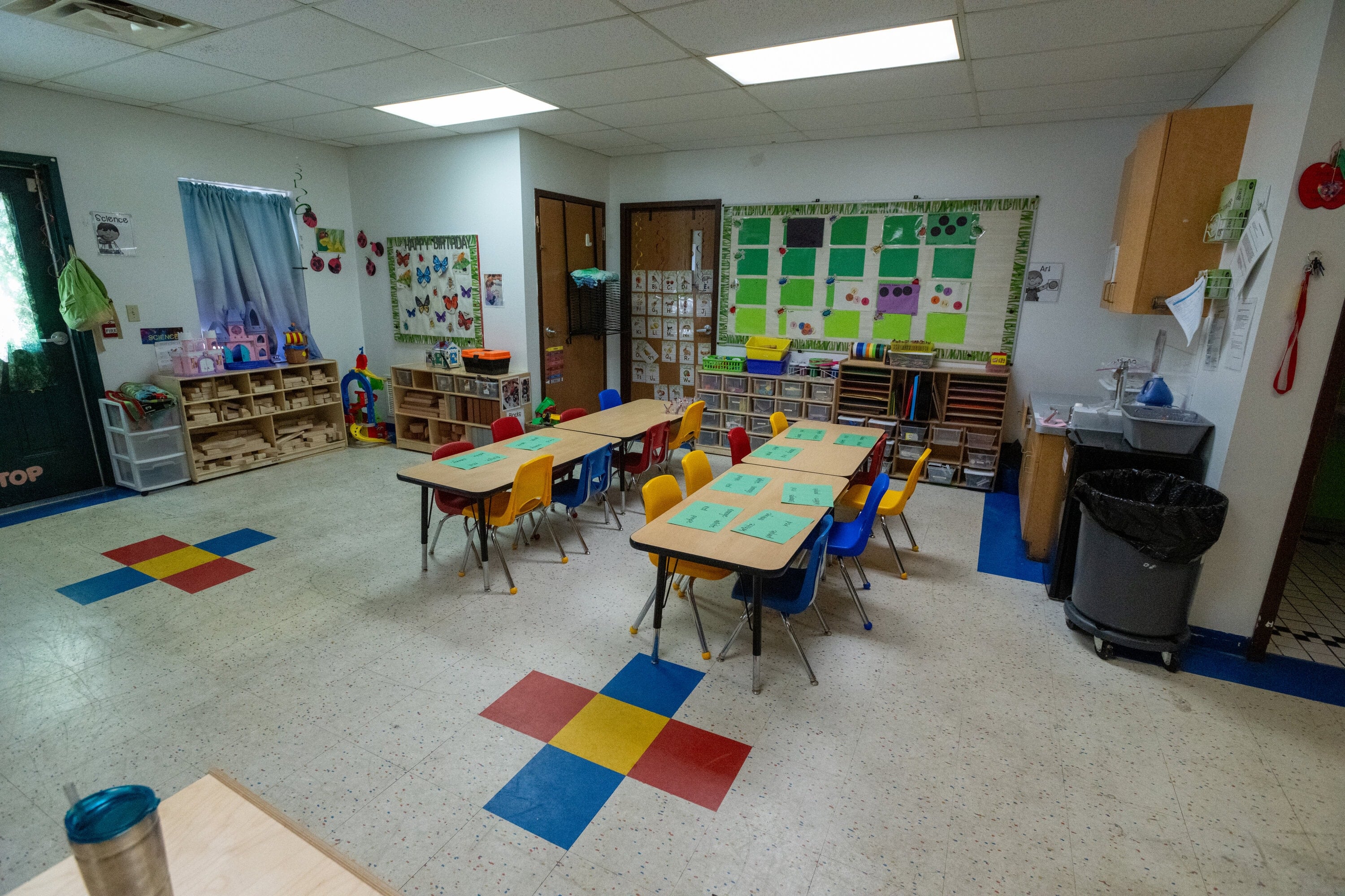 Classroom with small tables and colorful chairs arranged in groups, educational materials on the walls, and storage shelves in the background.