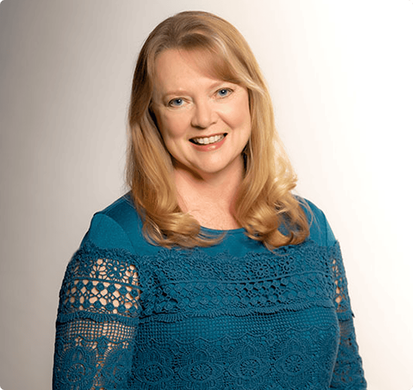 A woman with long blonde hair smiles at the camera, wearing a teal lace top against a plain light background.