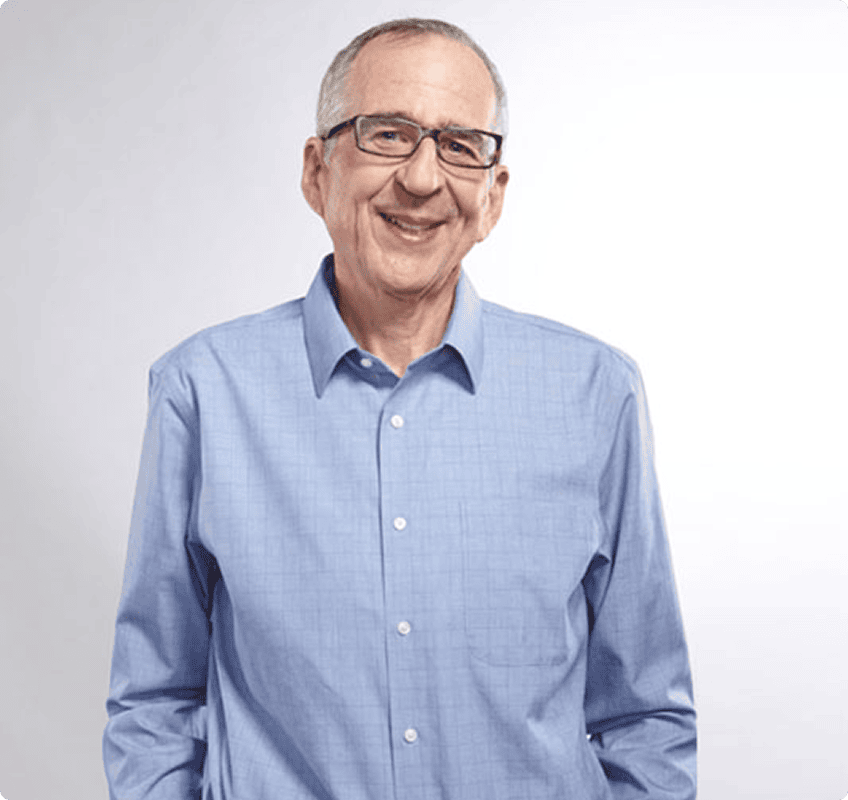 An older man with short gray hair and glasses stands against a plain white background, wearing a light blue button-up shirt and smiling.
