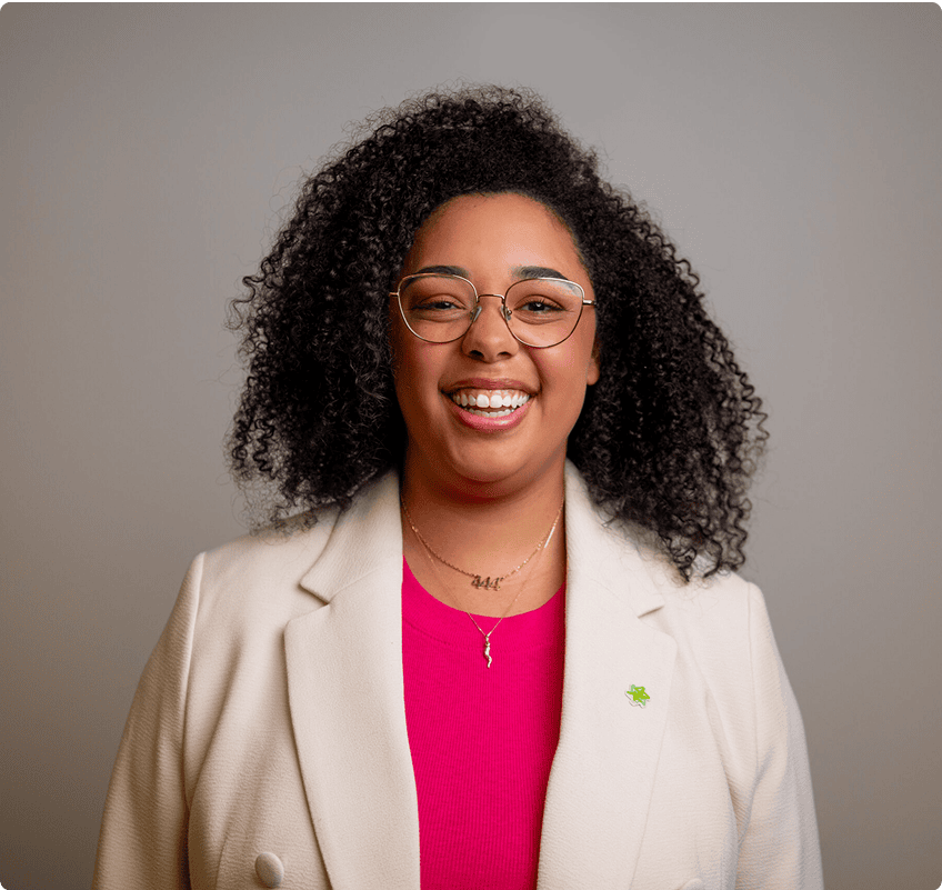 A woman with curly hair, wearing glasses, a white blazer, and a pink top, smiles in front of a plain gray background.