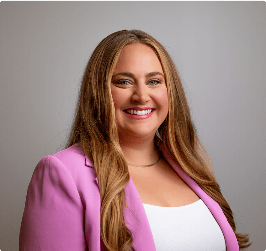 A woman with long, light brown hair wearing a pink blazer and white top smiles in front of a plain light background.