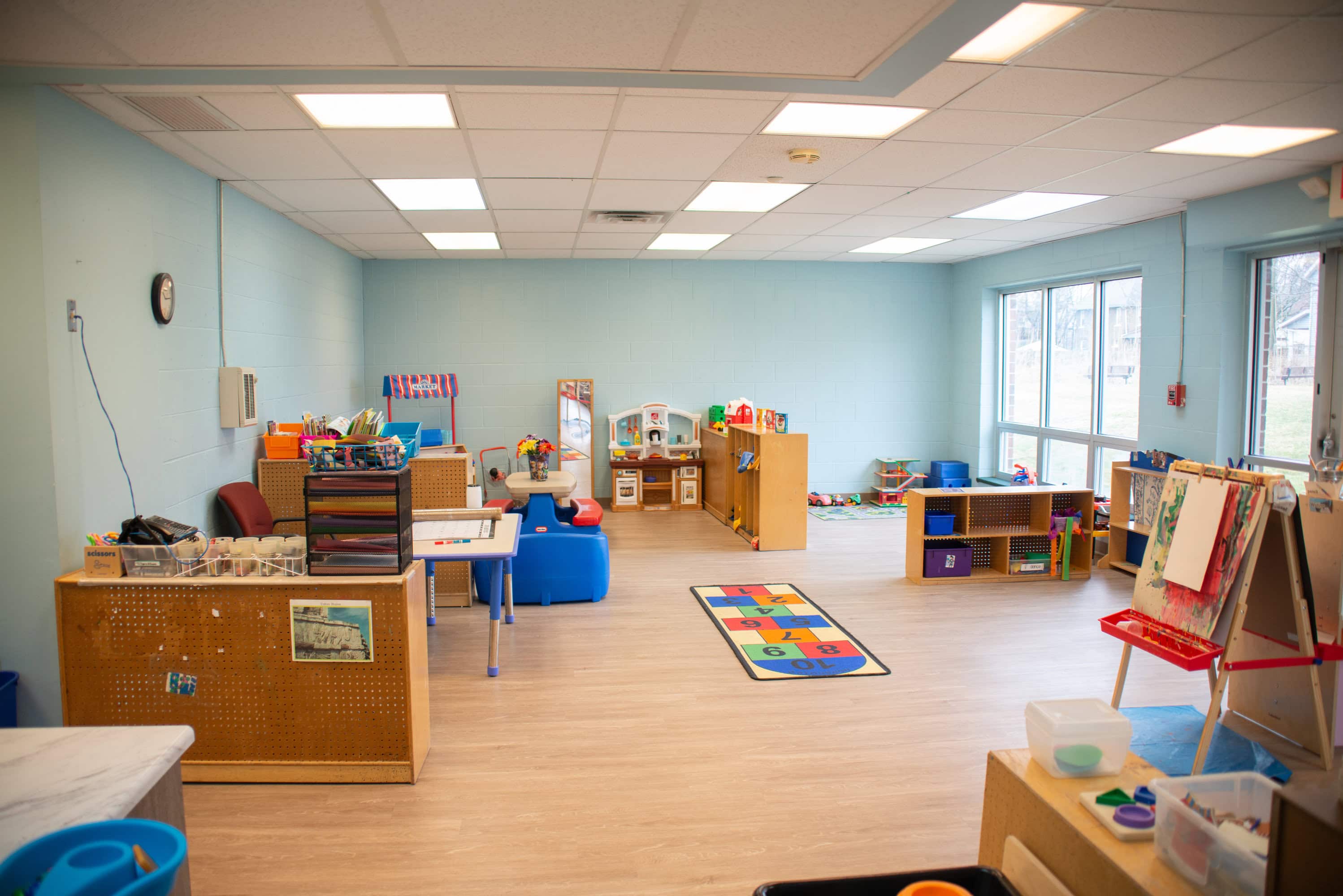 A brightly lit preschool classroom with organized play areas, toys, art supplies, a colorful rug, and large windows letting in natural light.