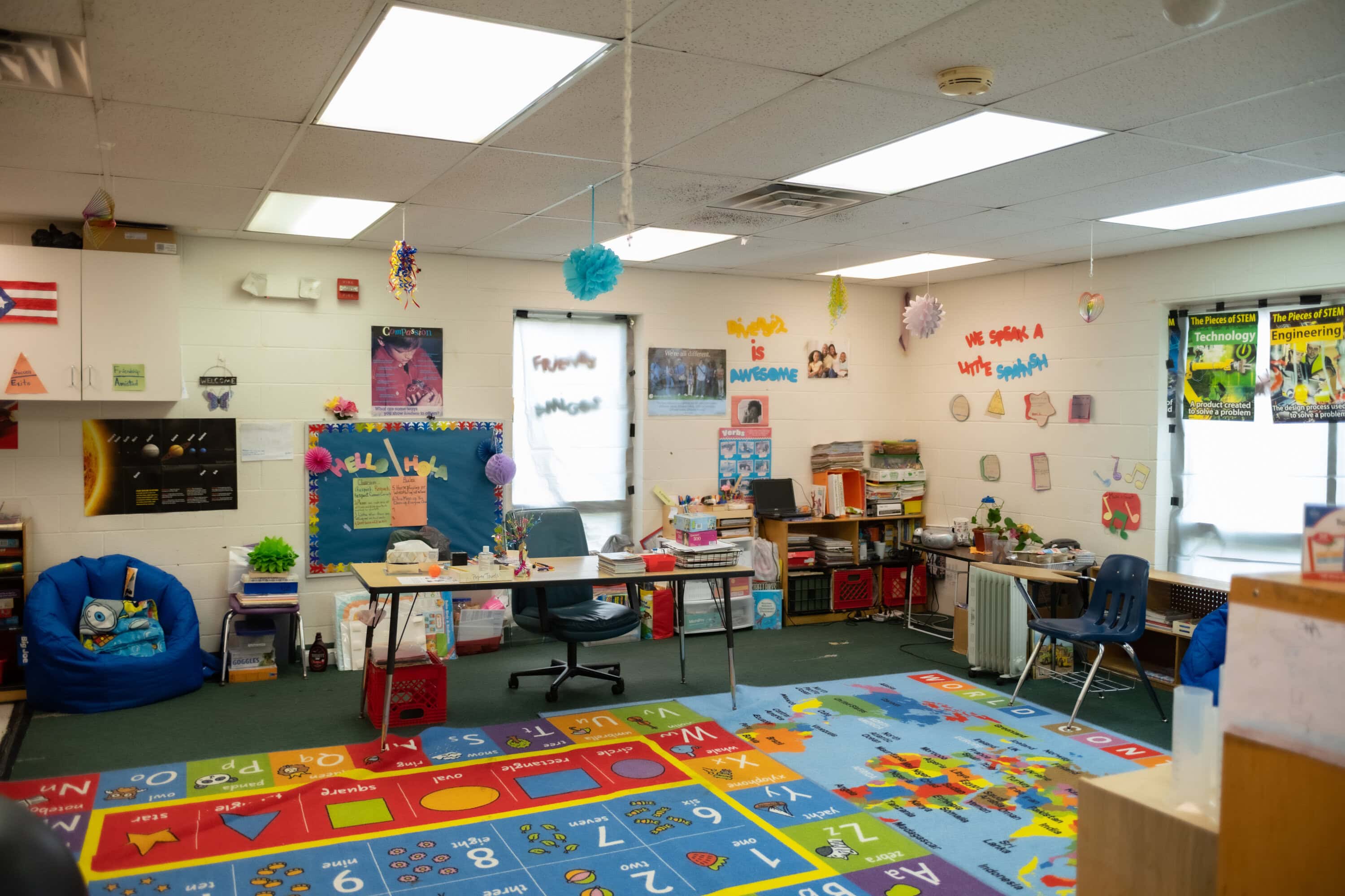 A colorful elementary classroom with alphabet and number rugs, student desks, educational posters on the walls, and various supplies neatly organized.
