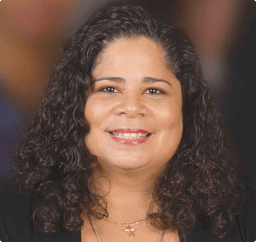 A woman with curly dark hair smiles at the camera, wearing a black top and a necklace with a gold pendant. The background is blurred.