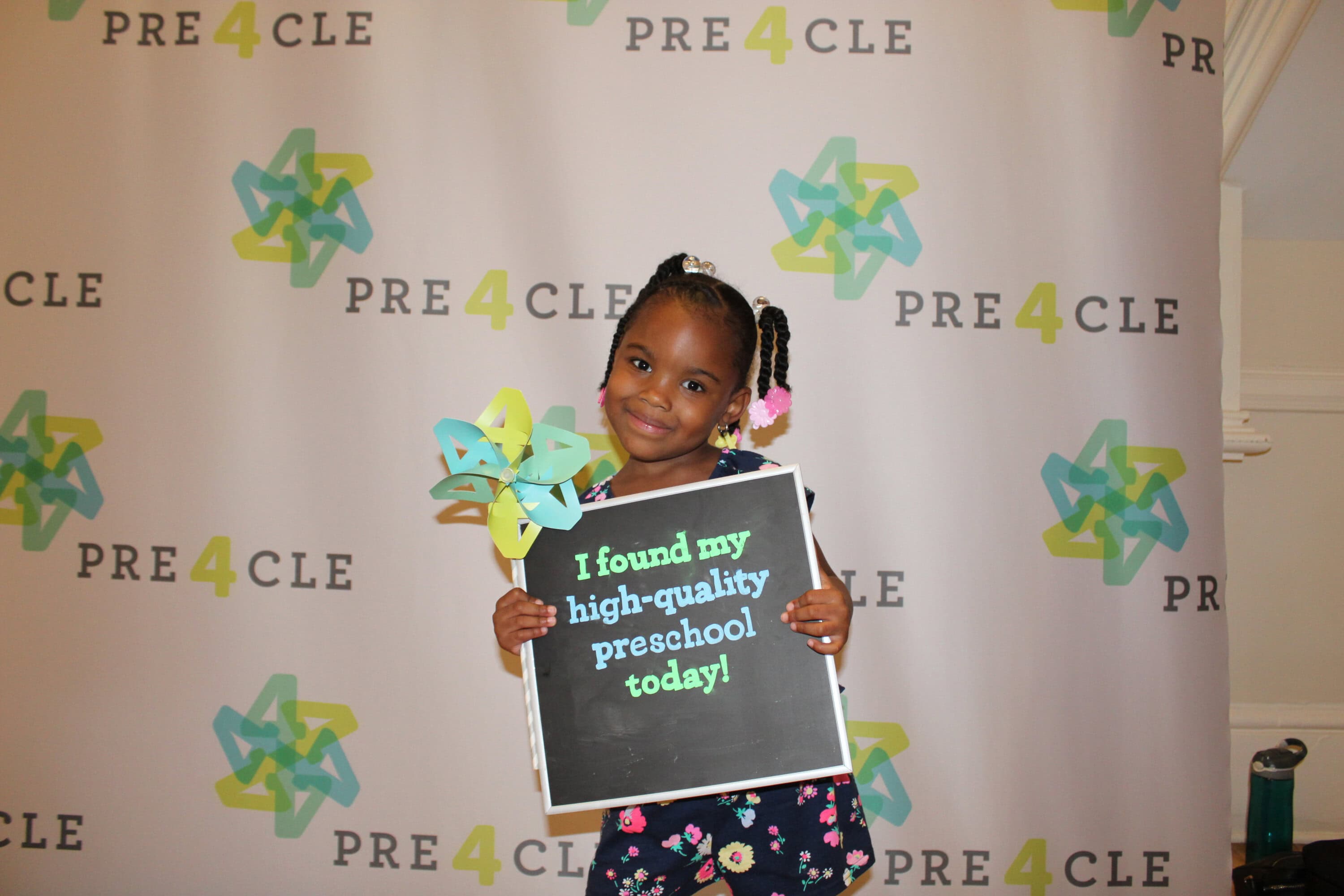 Young girl stands and smiles while holding a pinwheel and a sign that says, "I found my high-quality preschool today!" in front of a PRE4CLE step-and-repeat banner.