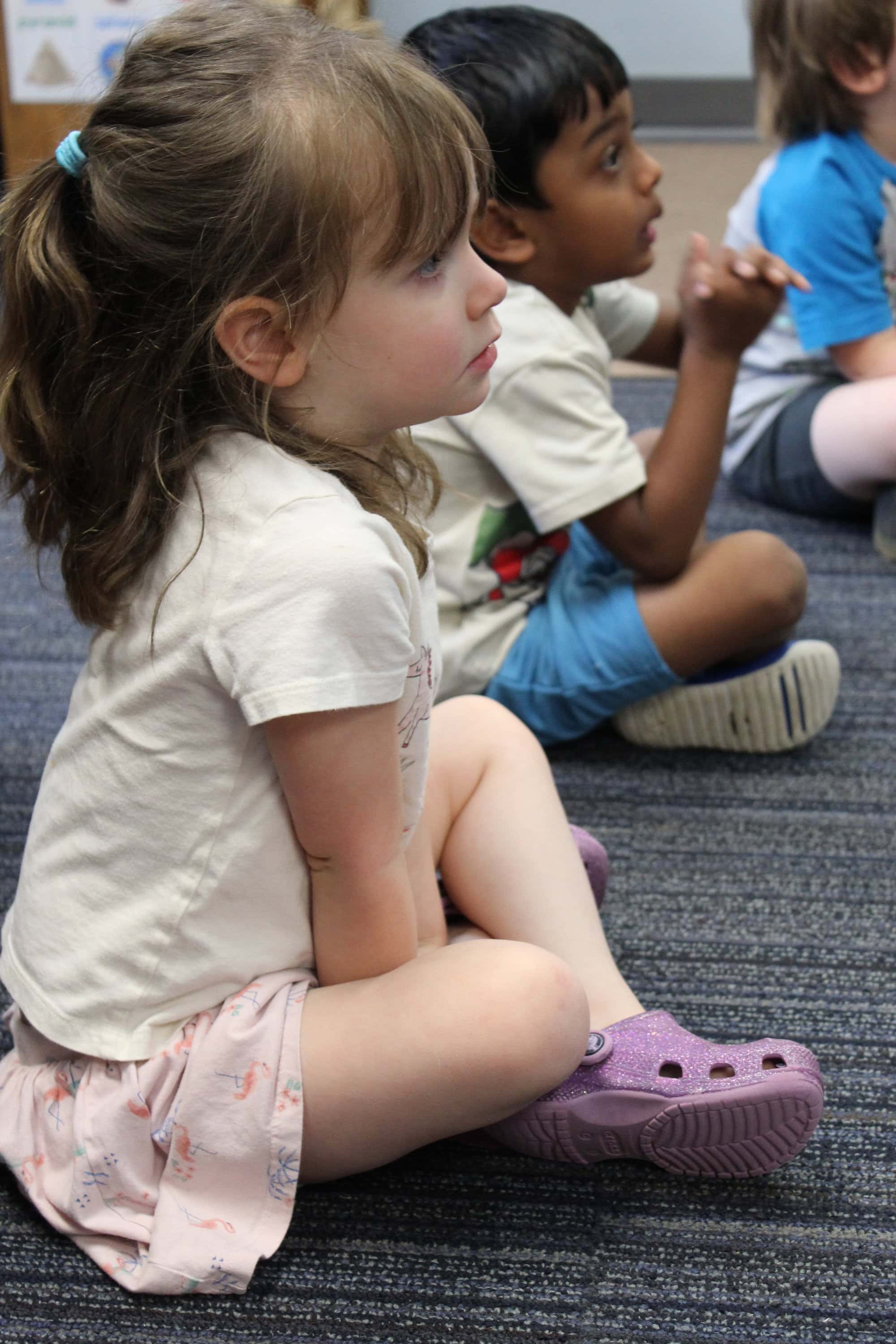 Young children sit cross-legged on a carpeted floor, attentively looking toward the front of a classroom.