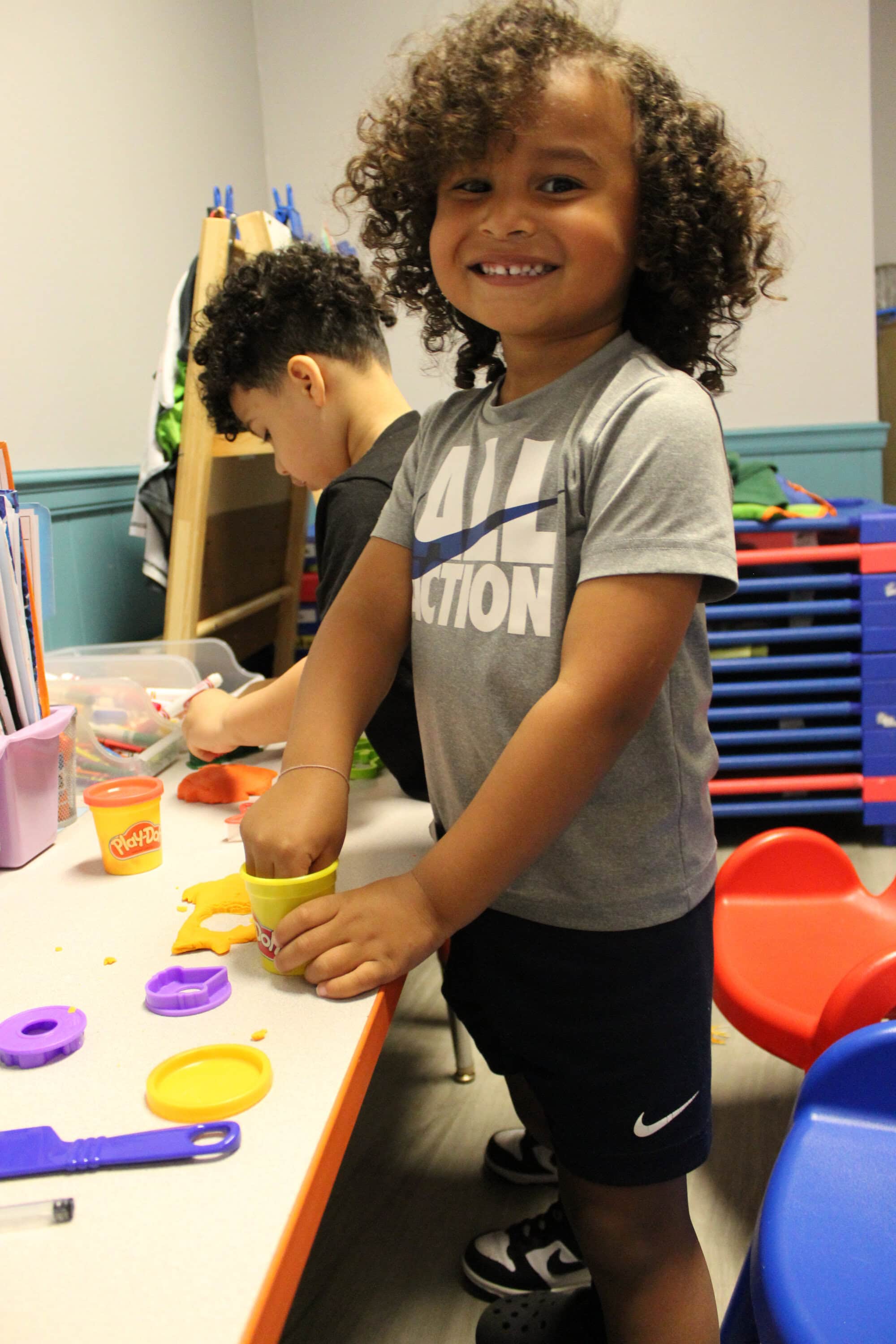 Two young children play with Play-Doh at a table in a classroom. One child is smiling at the camera while shaping dough; the other is focused on their activity.
