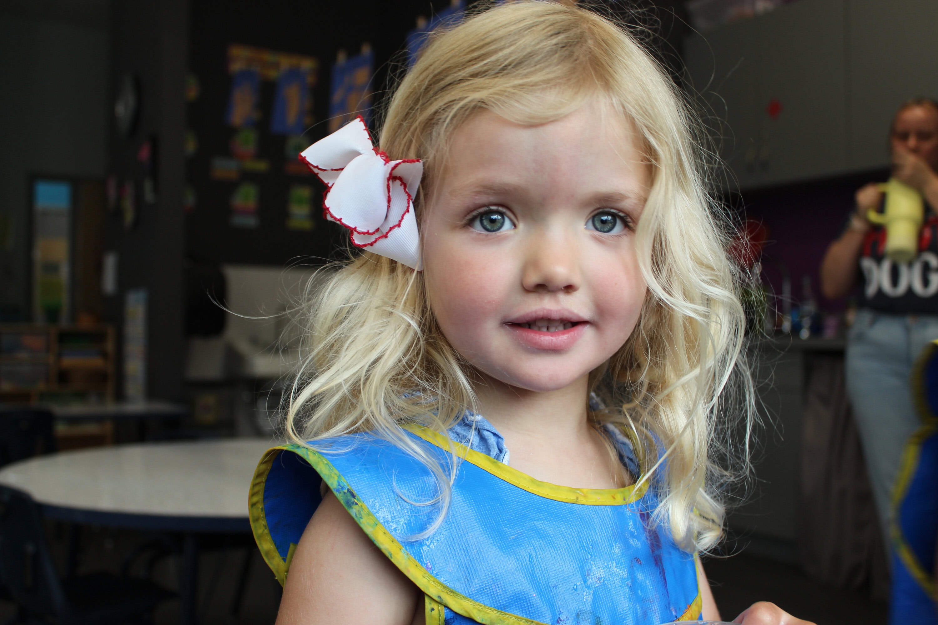 preschool-1 A young girl with long blonde hair and a white bow, wearing a blue smock, stands indoors in a classroom setting.