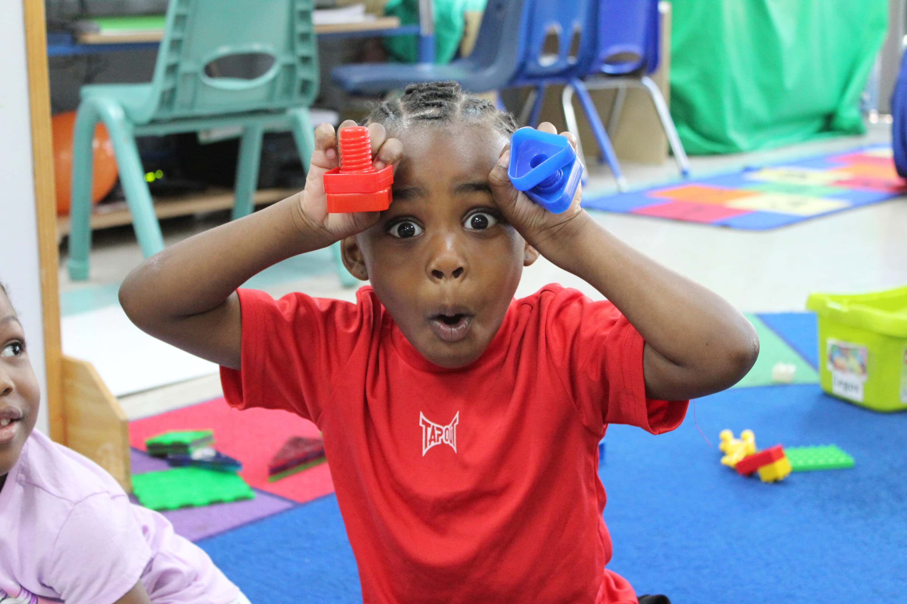 preschool-2 A young child in a red shirt holds colorful plastic toys to their face and makes a surprised expression in a classroom setting.