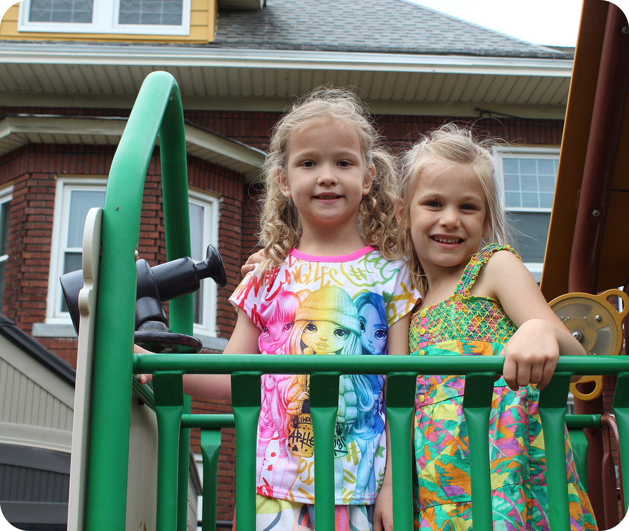 preschool-3 Two young girls stand side by side on a playground structure, smiling at the camera, with a brick house visible in the background.