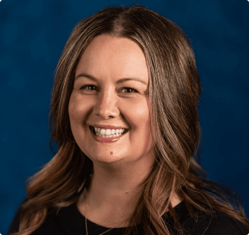 A woman with light brown hair smiles at the camera, wearing a black top against a solid blue background.