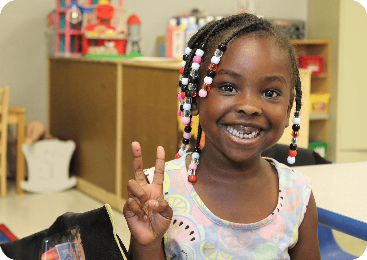 snapshot-2 A young girl with beaded braids smiles and flashes a peace sign while sitting in a classroom.