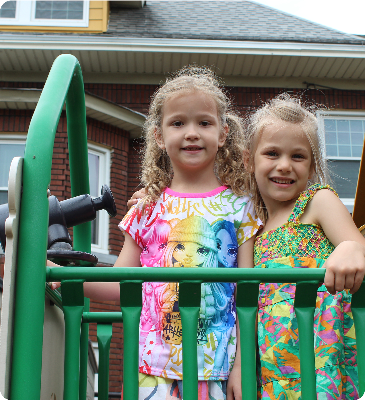 snapshot-3 Two young girls stand and smile on a green play structure outdoors, with a brick house visible in the background.