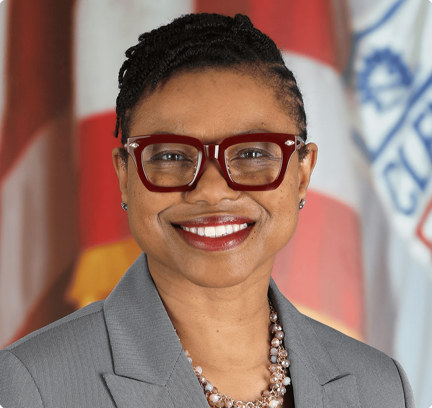 A person wearing glasses, a gray blazer, and a pearl necklace smiles in front of an American flag and a blue and white banner.
