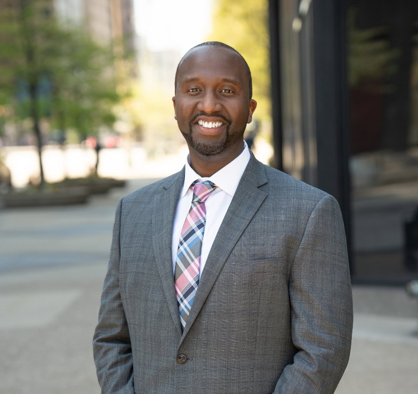 A man in a gray suit and plaid tie stands outdoors in a city setting, smiling at the camera. Trees and buildings are visible in the background.